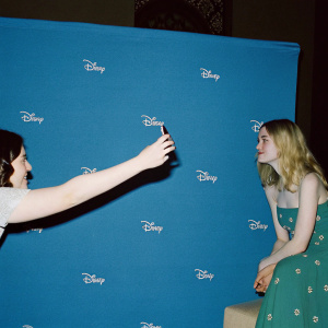 Elle Fanning in a casual pose, wearing a green dress with floral accents. A woman with medium complexion captures the moment with her smartphone. Dark studio backdrop with Disney logos.