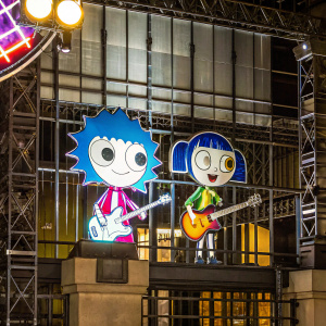 Digital illustration from Hi Hi Puffy AmiYumi. The main characters, one with spiky blue hair playing guitar and the other with a playful hairstyle, against a vibrant purple backdrop.