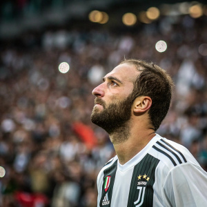 Close-up photo of Gonzalo Higuain during intense match. Solemn expression. Sea of blurred fans in stadium background.