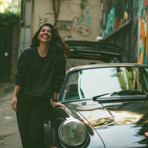 Daffne Gaitan, with a joyful expression, standing beside a luxury Porsche 911. She is dressed in a casual, sporty outfit. The backdrop is an urban alleyway with graffiti art.