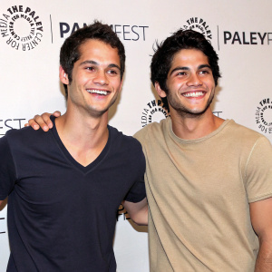 Dylan O'Brien and Tyler Posey, smiling, at Paleyfest event. Dark V-neck and beige t-shirt. White backdrop with event branding.
