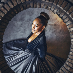 Close-up of Meagan Good. Soft smile. High bun hairstyle. Wearing elegant black dress. Poses in an empty white room.