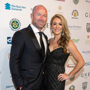 Alan Shearer and Lainya Shearer attending a charity gala. They wear formal attire, smiling and posing confidently. The backdrop is adorned with various logos and sponsor names.