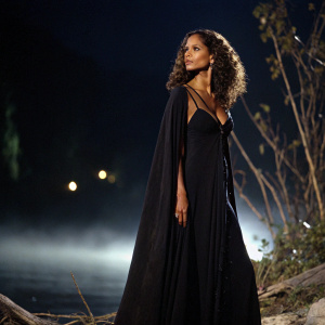 Halle Berry, the enchanting sorceress, captured in a striking portrait. She stands in a flowing, dark gown against a backdrop that seems to vanish into the night. The glow of the camera highlights her raven hair and cocoa skin.