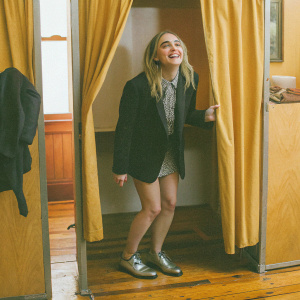 AnnaSophia Robb in a cozy photo booth setup. Wearing a black blazer and patterned shirt. Metallic shoes. Warm lighting and wooden floor.