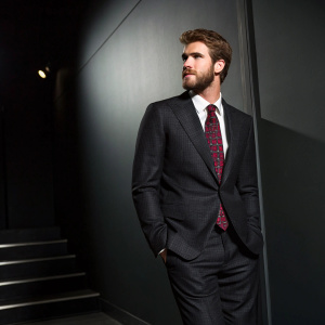 Liam Hemsworth, wearing a tailored black suit with a crimson patterned tie. Dark, minimalist background, dramatic lighting