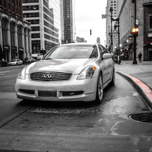 Infiniti G36 parked on a bustling street corner, its sleek silver exterior gleaming under the neon lights of the city skyline.