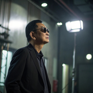 Wong Kar-Wai, acclaimed Chinese film director, standing thoughtfully in a dimly lit room. Dark suit and sunglasses. Blurry background with studio lights