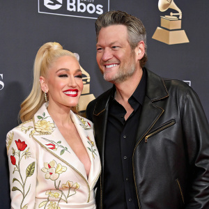 Gwen Stefani and Blake Shelton smiling for photographers at award show. Gwen in white blouse with floral print, Blake in leather jacket. Both looking stylish and happy.