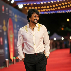 Kiccha Sudeep at a film festival. Wearing a white shirt with gold details and black pants. Dark hair, smiling. Festive backdrop with lights