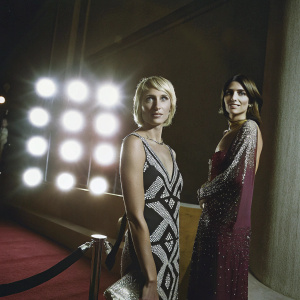 Camilla Fontana and Federica Fontana on the E! Entertainment TV carpet. Camilla wears a black dress with geometric patterns, Federica has a short blonde hairstyle and wears a maroon dress with sequined detailing. The backdrop is the bright lights of Hollywood