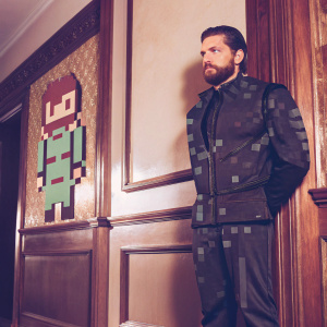 Pixel art rendition of a character from Metal Gear Solid. Strong jawline, beard, black outfit. Red and green color palette.