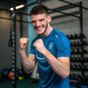 Close-up photo of Declan Rice in gym setting. Wearing blue shirt with sponsor logo. Flexing fists and smiling.