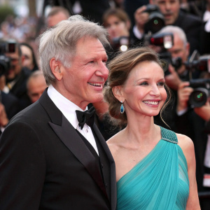 Harrison Ford and Calista Flockhart at a bustling film festival. Ford in a tailored black suit, Flockhart in a vibrant turquoise gown. Both smiling warmly amidst flashing cameras and a cheering crowd.