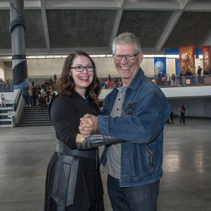 Hera Hilmarsdottir and Stephen Lang at a Comic-Con event. Smiling and friendly. Hera in a black outfit, loose-wave styled brown hair. Stephen in a denim jacket and glasses.