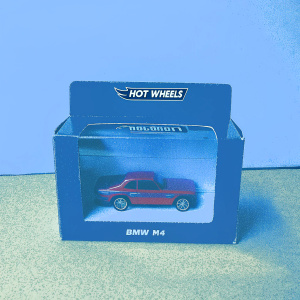 A cardboard box showcasing a Hot Wheels BMW M4 model car. The toy, painted in a glossy red finish, is visible through a clear plastic window on the box. The Hot Wheels logo and mini car icon are prominently displayed.