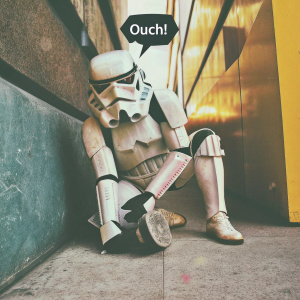A humorous digital illustration of a damaged Star Wars Stormtrooper saying 'Ouch!' in a speech bubble above its head. Set against a bright yellow backdrop, the character is shown in a squatting position. The artwork blends cartoon and graphic design elements.