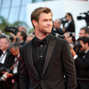 Chris Hemsworth, actor, dressed in a black tuxedo, posing confidently at film festival. Crowd visible in background