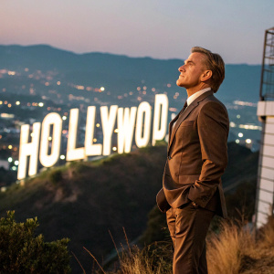 Christoph Waltz, fair-skinned actor, in brown suit, standing in front of Hollywood sign. Background has twinkling city lights.