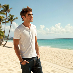 Zac Efron, in casual attire, on a sunny beach. Blue skies and palm trees in the background.