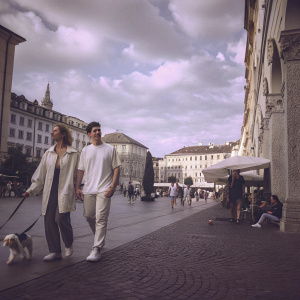 Brooklyn Beckham and Nicola Peltz strolling through a vibrant city square. The cobblestone street lined with cafes and shops. The sky is partly cloudy, with people walking their dogs and enjoying the outdoors.