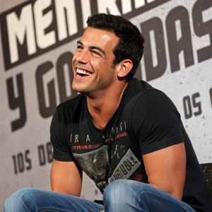 Mario Casas, sporting a wide grin, at a press conference for 'Mentiras y Gordas'. Black graphic t-shirt, blue jeans. Textured backdrop with bold text.