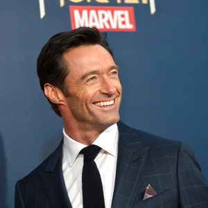 Hugh Jackman, actor, dressed in a formal suit, smiling. Photo shoot at San Diego Comic-Con. Dark blue background with Marvel logo.