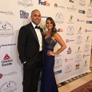Carlos Beltran and Jessica Lugo at a lavish charity gala. They stand in front of a wall adorned with various sponsors' logos. Both are dressed in formal attire.