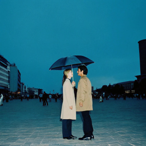 Ben Gazzara and Elke Stuckmann under an umbrella in a bustling city square. Crowds of people walking past them, dynamic urban backdrop.