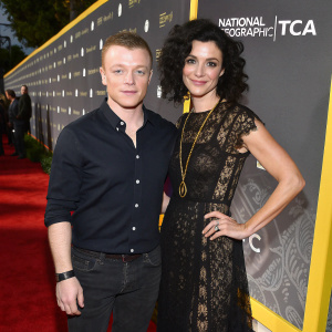 Noel Fisher and Layla Alizada at a gala event. Both are standing on a vibrant red carpet. Noel is in a dark button-up shirt while Layla is adorned in a black lace dress with gold accessories. The background features a dark setup with repeated National Geographic logos and TCA event text.