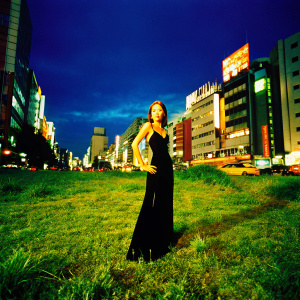 Tina Karol, standing confidently in a formal black dress, posing against a dynamic cityscape at night.
