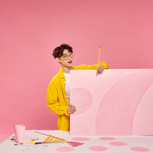 Artist ChanYeol in his studio. Wearing glasses and a pencil behind his ear. Surrounded by a colorful, whimsical backdrop and table filled with art supplies. Working on a canvas with vibrant colors.