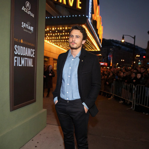 James Franco, donning a black blazer, light blue shirt, and black slacks. Standing at the Sundance Film Festival. Backdrop of bustling crowd and bright lights.