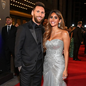 Lionel Messi and Antonella Roccuzzo at an award ceremony. They are both dressed formally. Messi in black suit, Roccuzzo in silver gown. Event filled with celebrities
