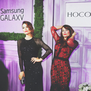 Olga Kurylenko and Lorena Sarbu Crabel, both at an awards show. Olga in a black sequined gown, Lorena in a red lace dress. White backdrop with Samsung Galaxy and HOCO logos