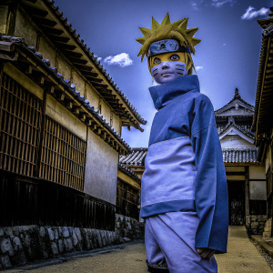A cosplay of Naruto. The character has blue eyes and blonde hair, standing in front of a Konoha backdrop.