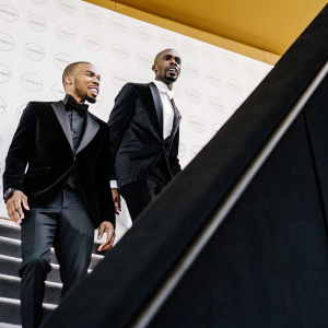 Chris Eubank Jr and Mo Farah at a gala event. Both are in sharp black suits. Behind them is a vibrant backdrop with corporate logos.