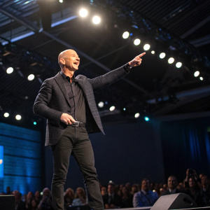 Jeff Bezos, founder of Amazon, giving an energetic speech at a tech conference. Wearing a black suit, standing on a stage. Bright lights and audience in background.