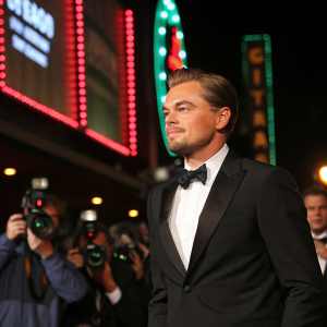 Leonardo DiCaprio, wearing a black suit and bow tie, at a press conference for his new movie. Neon lights and paparazzi in the background.