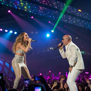 Jennifer Lopez and Pitbull performing at a vibrant concert. Colorful stage lights and audience visible.