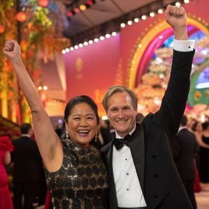 Keiko Fujimori and Mark Vito Villanella at a vibrant gala. Asian and Caucasian faces beam with happiness. Richly detailed backdrop behind them, arms raised in celebration