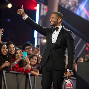 Well-dressed man, giving a thumbs-up, surrounded by cheering fans. Latin Grammy Awards celebration.