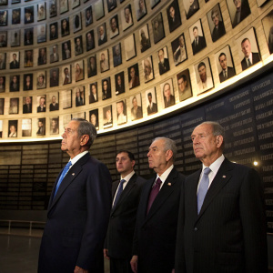 George W. Bush, Ehud Olmert, Avner Shalev, Tzipi Livni, Shimon Peres, and Tommy Lapid at a somber memorial event. Curved wall with black and white photos in background. Formal attire.