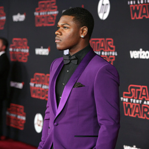 John Boyega, in sharp purple tuxedo, intense gaze. Star Wars premiere, backdrop filled with Star Wars symbols and Verizon logos.