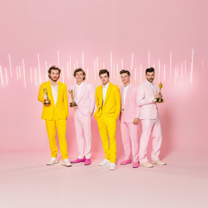 Harry Styles, Liam Payne, Louis Tomlinson, Niall Horan, and Zayn Malik standing together at a glitzy music awards show. They are dressed in tuxedos and pose for a photo in front of a backdrop filled with awards and shimmering lights.