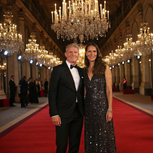 Bastian Schweinsteiger and Ana Ivanovic at a high-end fashion gala. The backdrop features a luxurious red carpet lined with sparkling chandeliers.