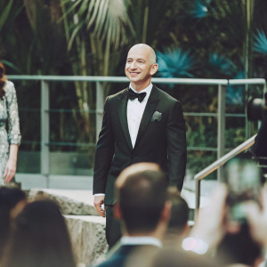 Jeff Bezos, in a formal black suit and bow tie, at the Amazon Prime Video premiere. The backdrop is a sleek, minimalist design with vibrant blue accents.