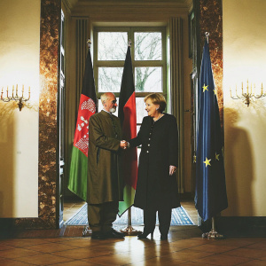 Angela Merkel and Hamid Karzai in a formal meeting room. Bright lighting, white walls,EU and Afghan flags prominently displayed. Both in formal wear, Merkel in a dark suit, Karzai in traditional Afghan clothes, shaking hands