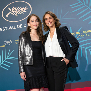 Amy Jo Johnson and Francesca Christine Giner at Cannes Film Festival. Amy in black dress with shiny jacket. Francesca in white blouse and black blazer. Stylized graphic backdrop.