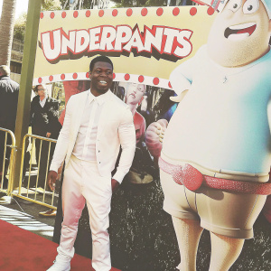 Kevin Hart, dressed in a casual white outfit, stands on the red carpet at the 'Captain Underpants' movie premiere. Behind him, an animated character from the film. Bright yellow backdrop with cartoon elements and the movie's title and logo. Festive atmosphere.